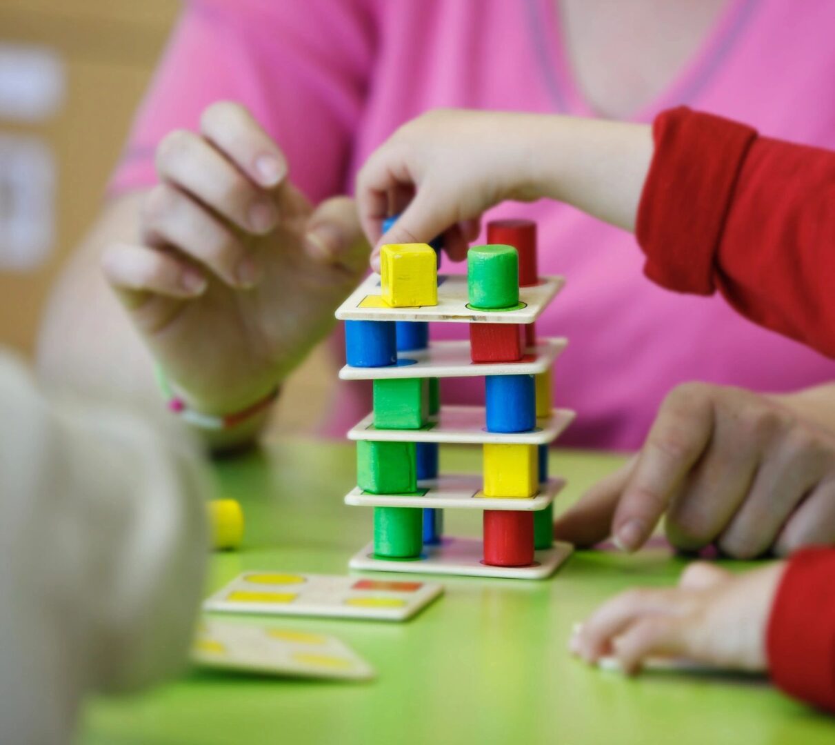 Children's hands stacking colorful wooden blocks on a green table, with an adult's hands assisting. This scene reflects the nurturing environment provided by Community Childcare Services.