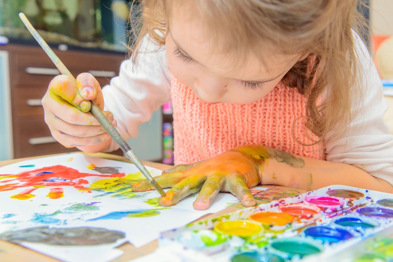 A child with paint on her hands and arm is using a brush to paint on a sheet of paper, with a watercolor palette nearby. This activity is often encouraged in early education programs to foster creativity and fine motor skills.