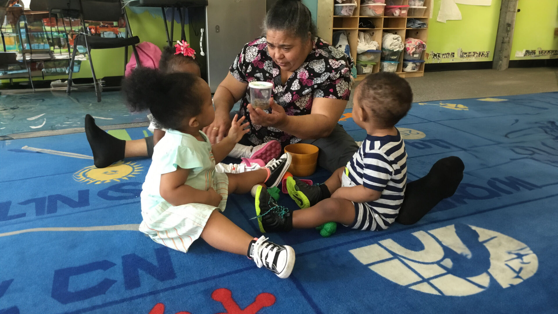 A woman engaging two toddlers with a puppet in a colorful classroom.