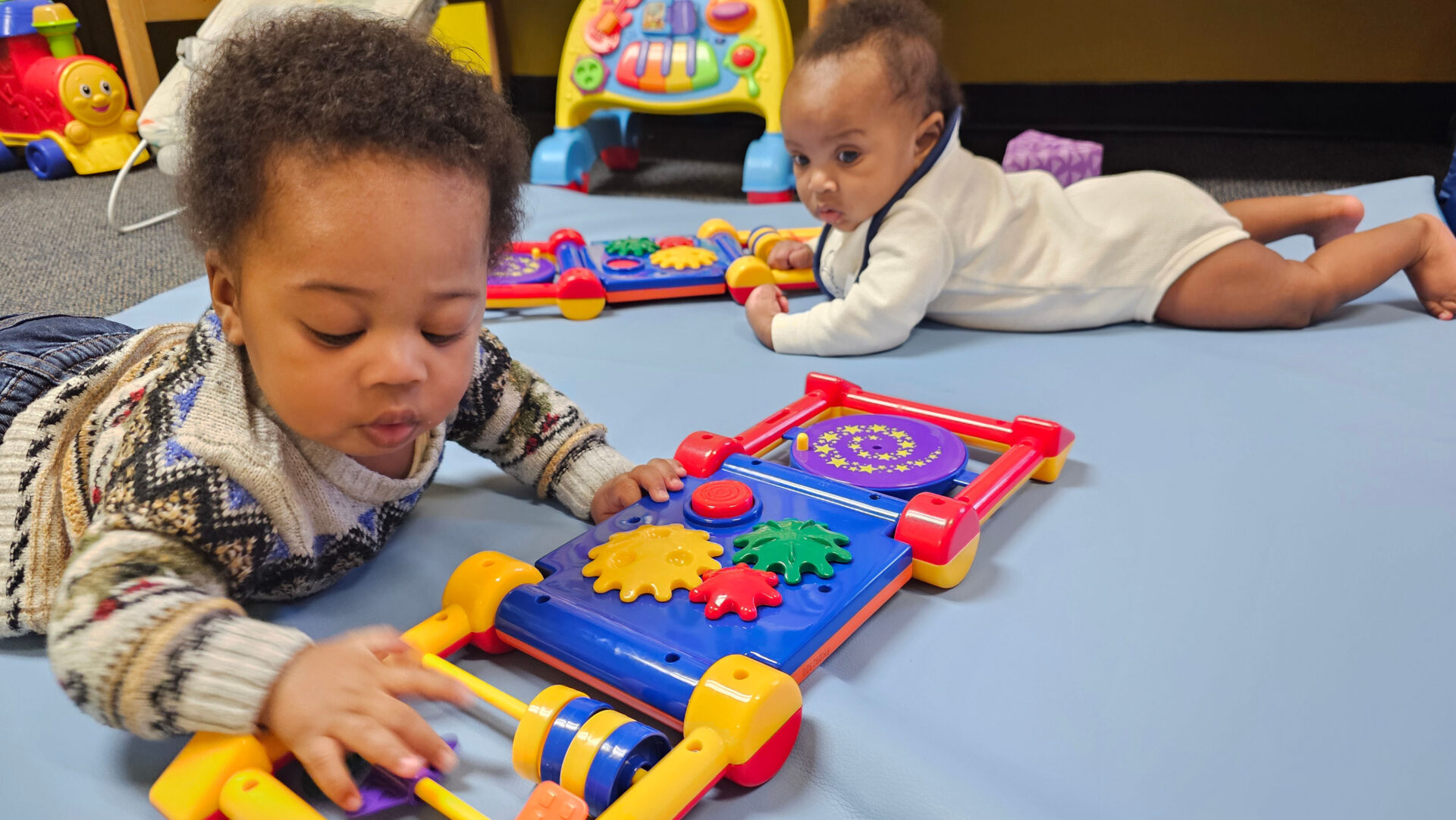 Two babies playing with colorful educational toys on a blue mat.